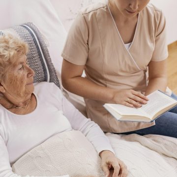 Helpful young carer reads a book to an elderly woman lying in a hospital bed